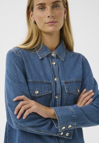 Keskmise sinise denimi särk klassikalise kraega, kuldsete nööbiga, kahe ees oleva tasku ja mansetidetailidega. Sujuv tekstuur ja mugav lõige.