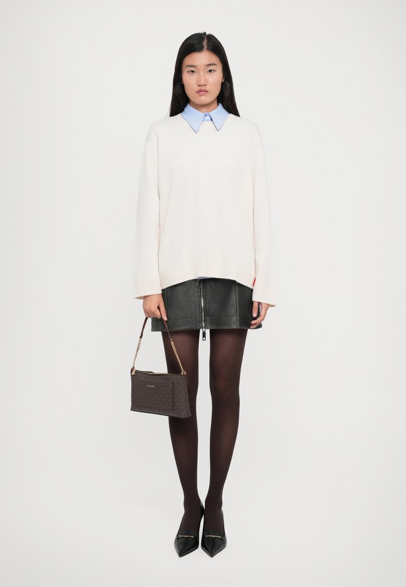 Femme portant un pull blanc sur une chemise à col bleu, une mini-jupe en cuir noire, des collants noirs, des talons noirs, tenant un sac à main en cuir marron à ses côtés.