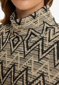 Beiger Pullover mit hohem Kragen, der geometrische schwarze und goldene Muster zeigt. Die Textur ist gestrickt mit einer Mischung aus glatten und erhabenen Fasern.