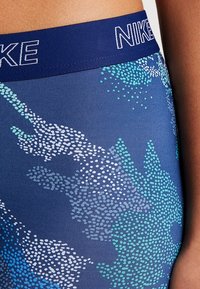 Detailní záběr na modré legíny Nike s tečkovaným abstraktním květinovým vzorem a námořnickým páskem s logem NIKE v bílé výšivce.