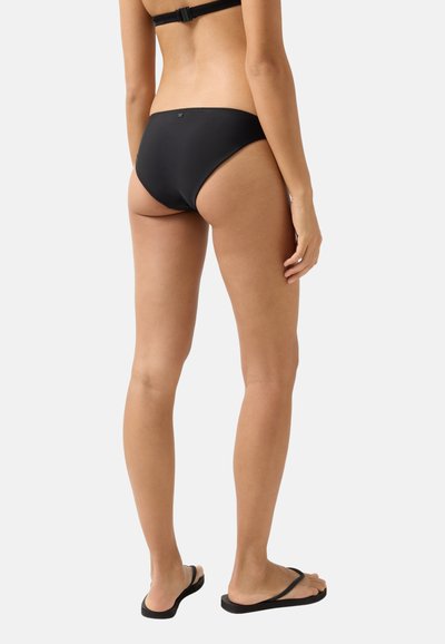 Frau mit schwarzem Bikinihöschen und Sandalen, von hinten vor einem schlichten hellen Hintergrund gezeigt.