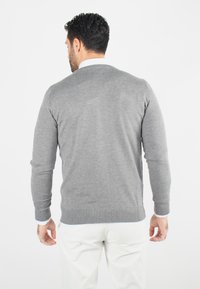 Pull en tricot gris avec de longues manches et un col rond. L'ourlet et les poignets sont côtelés, offrant une texture lisse. Porté sur un pantalon de couleur claire.