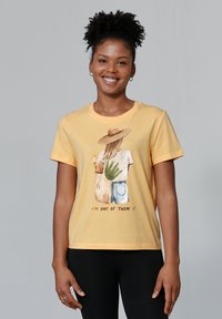 Gele katoenen t-shirt met een afbeelding van een persoon in een strohoed die planten en een baguette vasthoudt, met de tekst "IK BEN EEN VAN HEN" eronder.