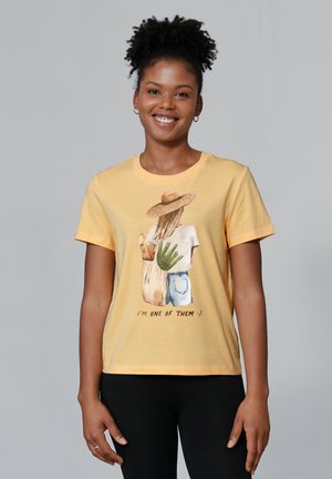 Gele katoenen t-shirt met een afbeelding van een persoon in een strohoed die planten en een baguette vasthoudt, met de tekst "IK BEN EEN VAN HEN" eronder.