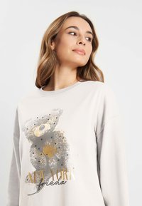 Lichtgrijze sweatshirt met een ronde hals, voorzien van een berenafbeelding in goud en grijs, versierd met zwarte studs en de tekst "NEW YORK".