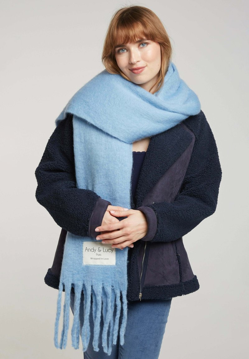 Écharpe en laine douce de couleur bleu clair ornée de bords à franges, portée sur une veste en polaire bleu marine foncé. Étiquette visible : "Andy & Lucy, Enveloppé d'Amour."
