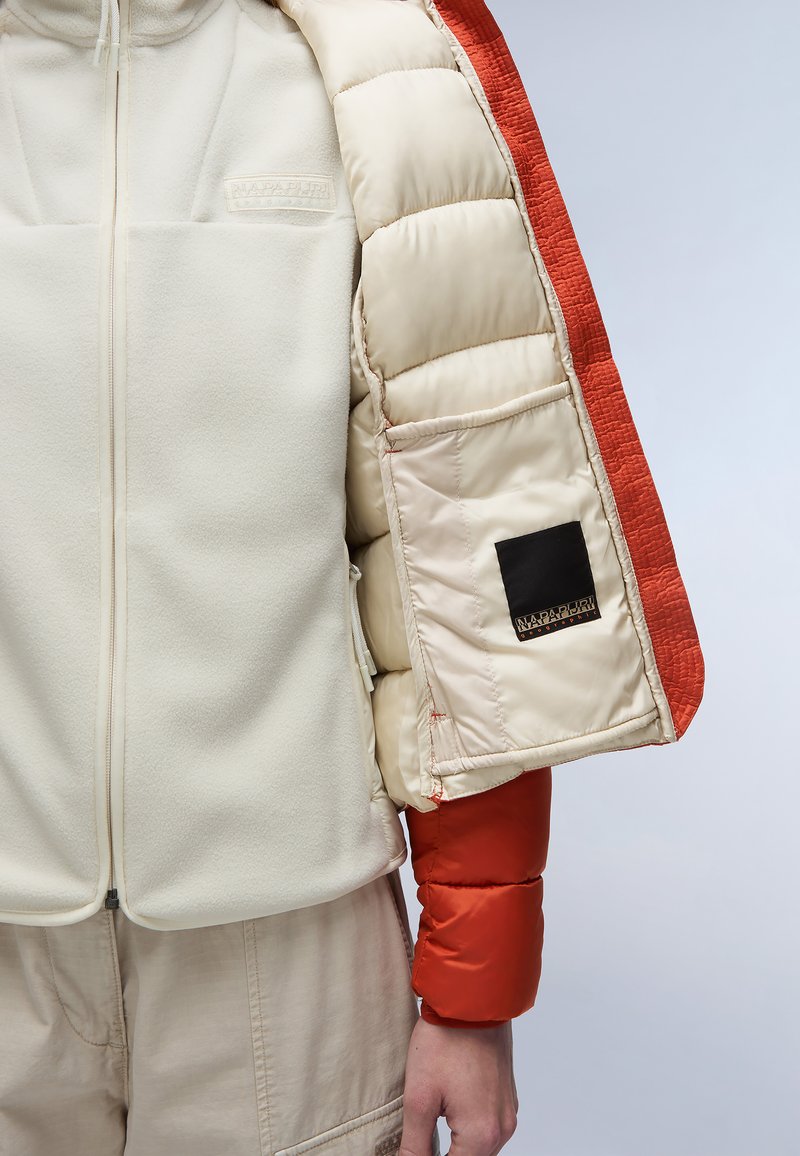 Giacca imbottita con corpo in pile crema, maniche arancioni e un'etichetta nera. Materiali testurizzati e modelli trapuntati visibili.