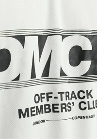 Bílá síťová látka se silným černým textem, který zní „OMC OFF-TRACK MEMBERS’ CLUB“ s nápisy „LONDÝN“ a „KOPENHÁGA“ pod ním.