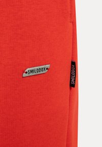 Czerwony materiał sportowy z teksturowaną powierzchnią. Posiada srebrną metkę z logo "SMILODOX" oraz czarną metkę marki na szwie bocznym.