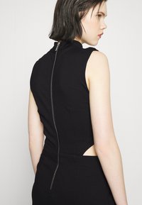 Vestido preto sem mangas com collar alto, apresentando um fecho de correr visível nas costas e detalhes recortados nos lados. Tecido suave e elástico.