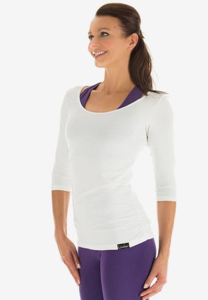 White long-sleeve top with a scoop neckline, paired with purple leggings. The top features a fitted design and slight ruching at the sides.