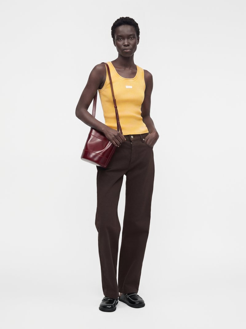 Femme portant un haut jaune sans manches, un pantalon marron foncé, des mocassins noirs et un sac à main bordeaux, debout sur un fond uni.
