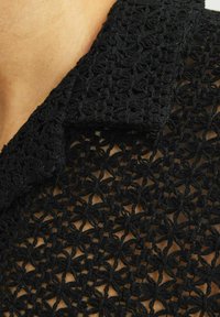Czarna, misternie szydełkowana tkanina z kwiatowymi wzorami oraz kołnierzykowym dekoltem, ukazująca fakturę i detale projektu z otwartym szyciem.