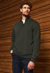 Tmavě zelený pulovr s čtvrtinovým zipem vyrobený z texturované látky, s malým logem na hrudi, žebrovanými manžetami a volným střihem.