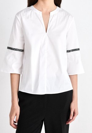 Vrouw draagt een witte blouse met V-hals en driekwart wijde mouwen met een zwart patroon als rand, gecombineerd met een zwarte broek.
