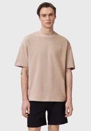 Man draagt een beige oversized T-shirt met korte mouwen met een klein rond logo op de borst en zwarte knielange shorts, staand voor een effen achtergrond.