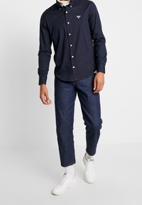 Laivastonsininen nappipaita valkoisilla napeilla, päällä tummat farkut ja valkoiset tennarit, seisomassa yksiväristä taustaa vasten.