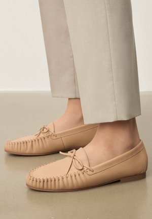 Mocassins en cuir beige avec détail nœud, portés avec un pantalon beige clair coupé court sur un sol lisse.