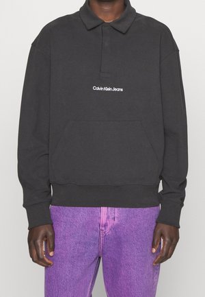 Osoba ubrana w czarny, długi rękaw polo Calvin Klein Jeans oraz fioletowe dżinsy, stojąca na jednolitym tle.