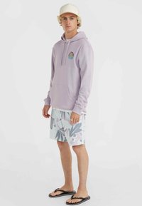 Lilac hoodie with front pocket and logo patch, paired with patterned pastel swim shorts featuring leaf designs. Black flip-flops complete the look.
