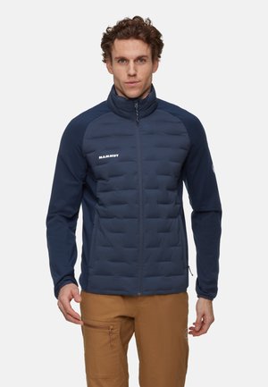 Giacca isolante blu navy con parte anteriore trapuntata, maniche lisce, colletto alto e sottile logo bianco. Chiusura frontale con cerniera e tasche laterali.