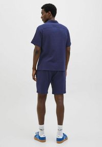 Chemise à col bleu marine à manches courtes, assortie à un short en tissu texturé. L'ensemble est complété par des baskets bleues.