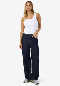 Mlada žena s dugom plavom kosom nosi bijelu majicu bez rukava, tamne široke traperice i srebrne sneakersice, stojeći nasuprot jednostavne pozadine.