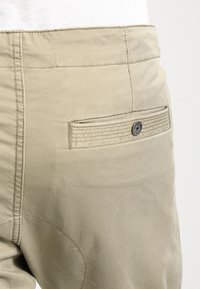 Primo piano di una tasca di pantaloni beige con pattina a bottoni e cuciture visibili su un tessuto liscio.