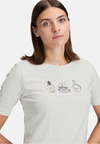 Jeune femme avec des cheveux bruns raides portant un t-shirt blanc arborant trois flacons de parfum étiquetés amour, rêves et espoir avec le texte "inoubliable".