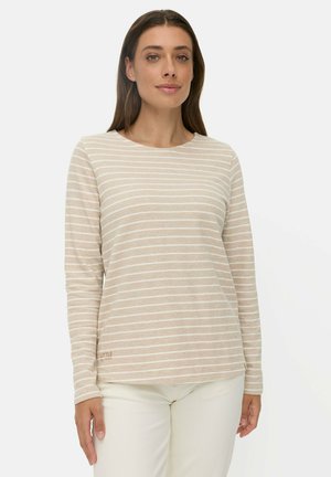 Frau mit langen braunen Haaren trägt ein beige-weiß gestreiftes Langarmshirt und weiße Hose, steht vor einem schlichten hellen Hintergrund.