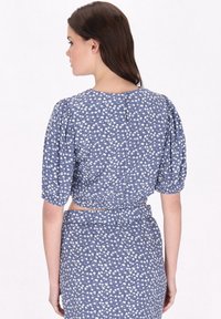 Top corto floreale blu con maniche a sbuffo e scollatura alta, caratterizzato da fiori bianchi su un tessuto a fantasia e apertura sulla schiena ben visibile.