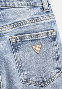 Jeans di denim azzurro chiaro con un logo triangolare "GUESS?" sulla tasca posteriore. Cuciture visibili in giallo e un dettaglio di tessuto testurizzato.