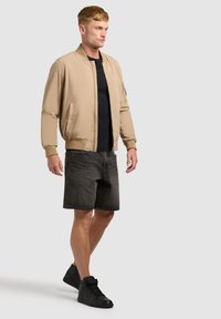 Homme debout portant une veste bomber beige, un t-shirt noir, un short en jean gris foncé et des baskets montantes noires sur un fond clair uni.