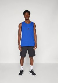 Μπλε αμάνικο με λογότυπο, μαύρο σορτς, μαύρα αθλητικά παπούτσια και λευκές κάλτσες. Το σύνολο είναι σχεδιασμένο για ενεργή αθλητική χρήση.