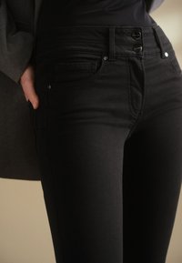 Gros plan sur une personne portant un jean noir taille haute avec fermeture à double bouton et la main glissée dans la poche de la veste.