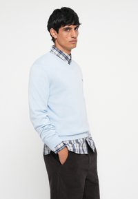 Lichtblauwe v-hals trui over een blauw-wit geruit overhemd, gecombineerd met donkere corduroypantalon. Bevat ribgebreide manchetten en tailleband.