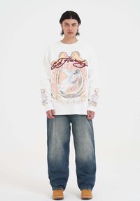 Witte sweatshirt met kleurrijke grafische ontwerpen van een paard en vlammen, gecombineerd met losse blauwe jeans en gele klompen.