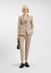 Beige pak met een getailleerde blazer en op maat gemaakte broeken, voorzien van subtiele zakdetails, gecombineerd met stevige zwarte loafers en een kleine handtas.