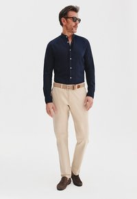 Granatowa lniana koszula z guzikami, beżowe spodnie, brązowy pasek i ciemne buty. Model stoi w okularach przeciwsłonecznych na tle jasnym.