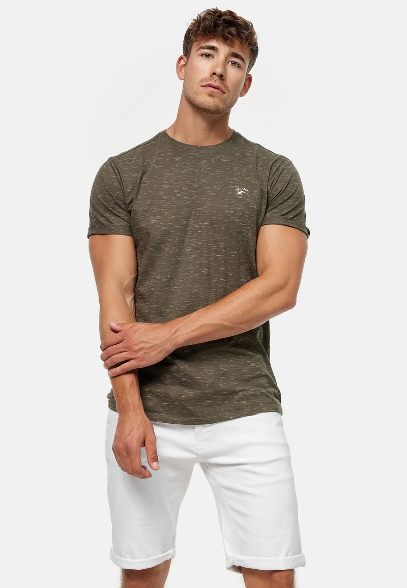 Olivgrünes T-Shirt mit kurzen Ärmeln und subtiler Textur sowie einem kleinen Logo, kombiniert mit weißen, hochgekrempelten Jeansshorts.