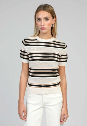 Jeune femme aux cheveux blonds raides portant des boucles d'oreilles créoles, un haut en maille crème à manches courtes avec des rayures horizontales noires et marron, et un pantalon blanc.