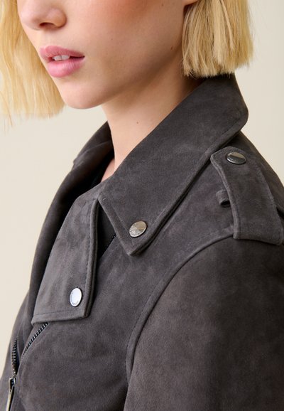 Giacca in suede grigia con colletto strutturato, bottoni a pressione e chiusura con zip; presenta una texture liscia e una vestibilità su misura.