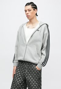 Pessoa jovem com cabelo trançado, vestindo um casaco cinzento da Adidas com fecho éclair e riscas pretas, e calças estampadas com logótipos da Adidas.