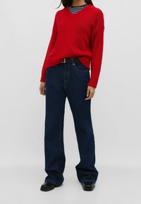 Rode gebreide trui met een v-hals en geribbelde manchetten, gecombineerd met donkerblauwe wijde jeans. Zwarte riem met een gouden gesp.