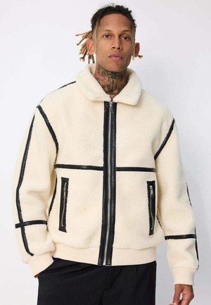 Homme aux dreadlocks portant une veste en polaire crème avec des bordures noires et des poches zippées, debout devant un fond blanc uni.