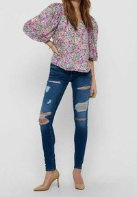 Blumenoberteil mit Puffärmeln in Pink, Lila und Grün; kombiniert mit blauen, gerissenen Skinny Jeans; hautfarbene, hochhackige Schuhe.