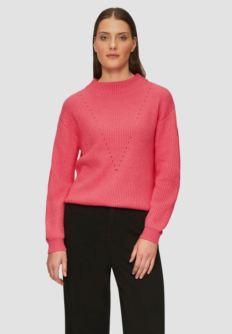 Helle pinke Rippstrick-Pullover mit hohem Ausschnitt, der ein V-Muster und Diamantakzente aufweist. Kombiniert mit schwarzen Hosen für den Kontrast.