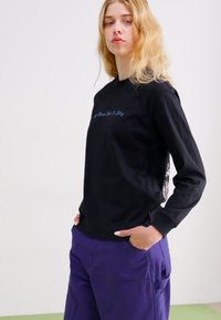 Crna majica dugih rukava s izvezеним plavim tekstom sprijeda. Ima bočni panel s uzorkom i ležeran kroj. Uparena s ljubičastim hlačama.