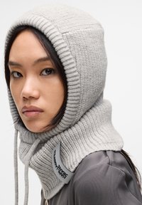 Scarf a cappuccio lavorato a maglia grigio chiaro, con texture a coste. Include un cordoncino a loop e un'etichetta nera con logo per un contrasto distintivo.