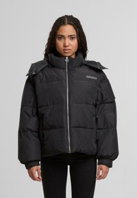 Giovane donna con capelli intrecciati che indossa un giubbotto trapuntato nero con colletto alto e zip frontale, in piedi di fronte a uno sfondo grigio chiaro uniformemente.